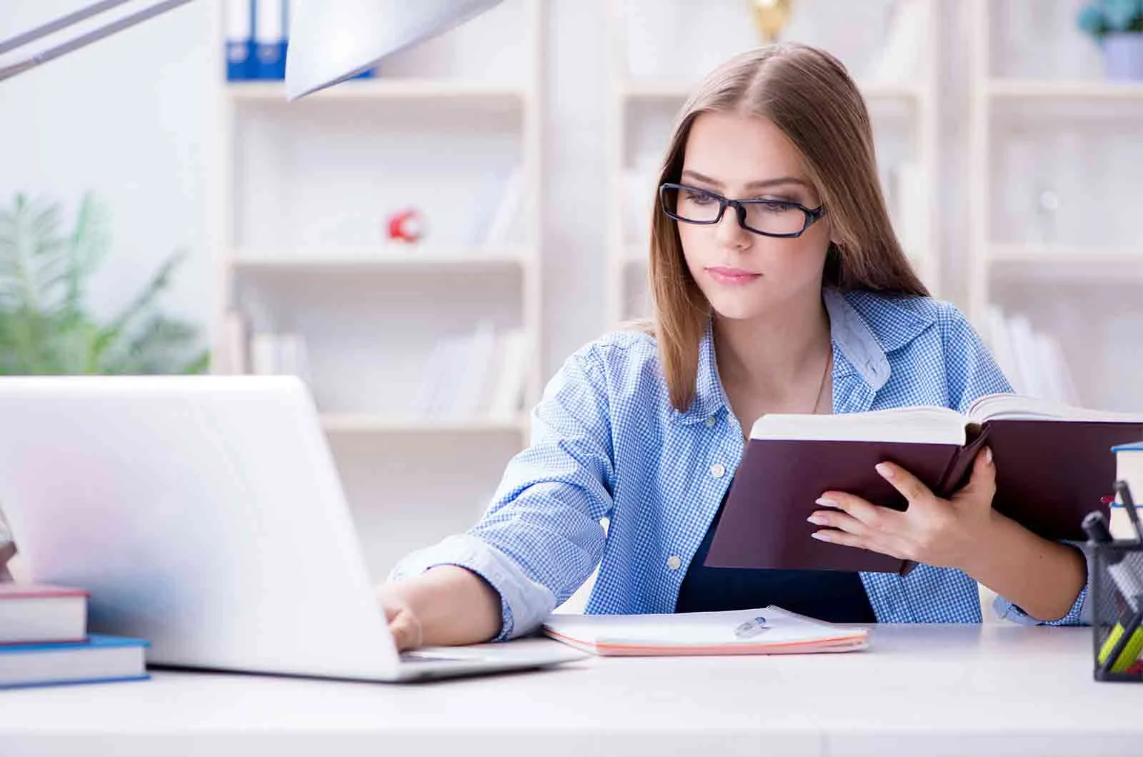 A girl wearing glasses and holding a book while using a laptop. Concept of online editing and proofreading services.