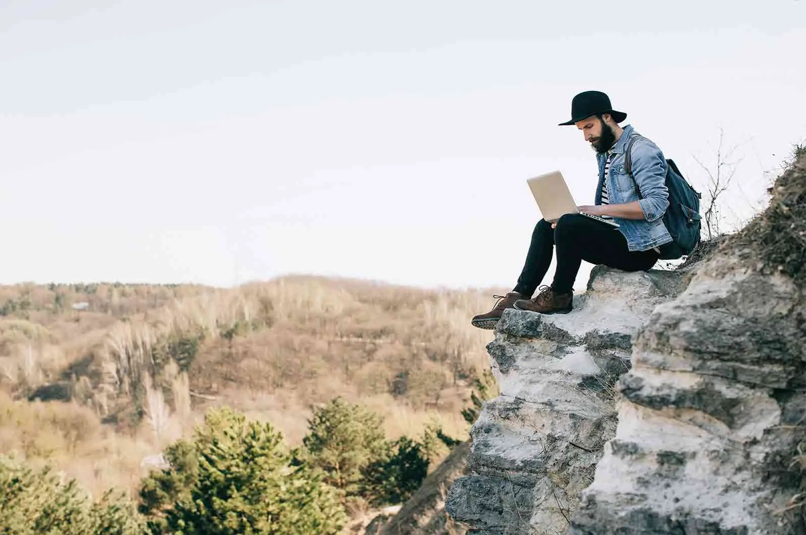 A man wearing a backpack sits on a cliff using his laptop. Concept of freelance translators working for translation agencies.