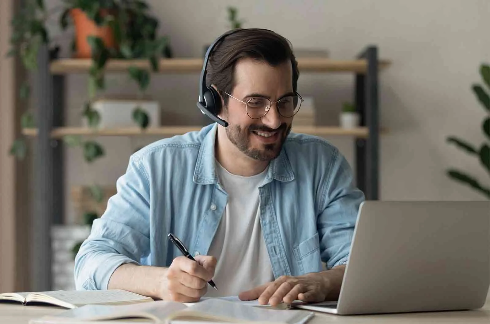 A man with headphones smiling, taking notes and using laptop. Concept of language skills and development.