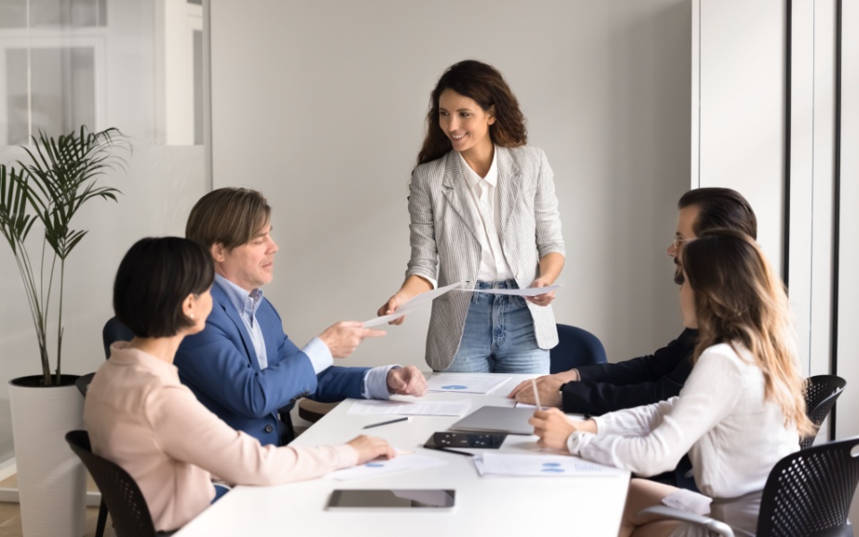 During a fruitful staff training meeting in the conference room, a cheerful young lady corporate trainer office coach shared printed handout materials with trainees from a variety of company groups; training materials transla