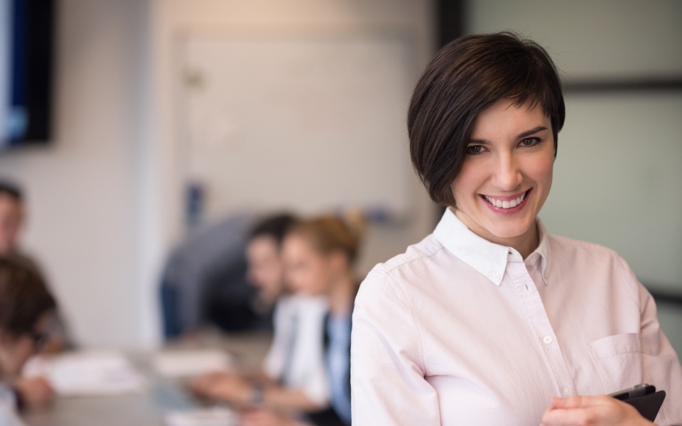 Young businesswoman with tablet computer at modern business office with people meeting in background discussing cheap translation services. 