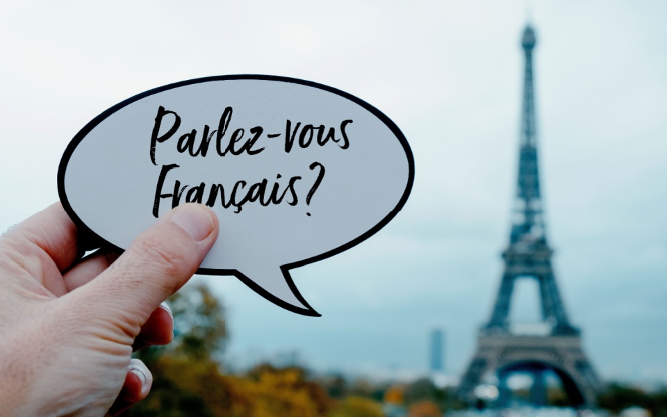 In front of the Eiffel Tower in Paris, France, a young man is holding a speaking balloon that reads, "Do you speak French accents?" printed in the French language. 