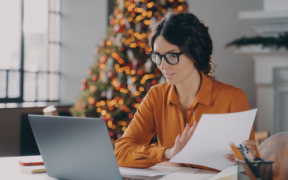 Hispanic lady wearing black glasses and working as translation project manager in office, sitting at desk and working on computer with papers in hands. 