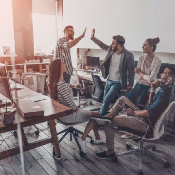 Two young employees celebrate success by giving high-five while their colleagues sitting and smiling. 