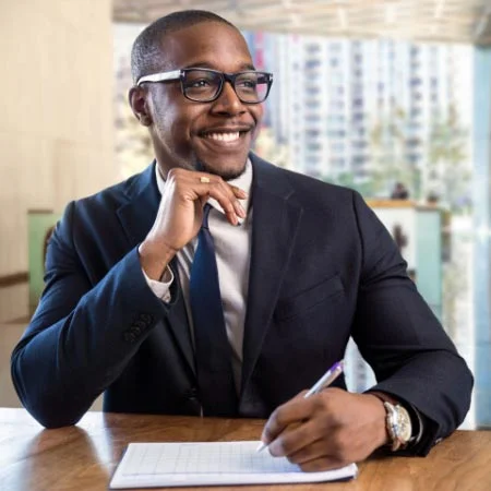 African man wearing suit and glasses smiling and signing paper.