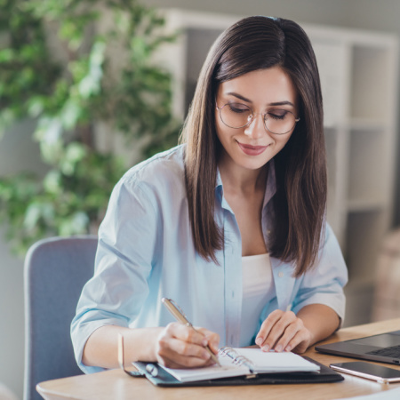 Pretty young woman working on her desk using laptop and taking notes on paper. Concept of professional proofreaders.