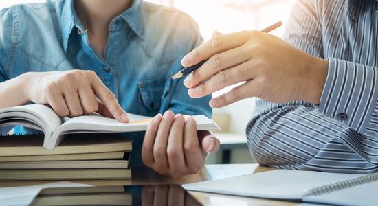 One person holding an open a book, the other pointing at it. 