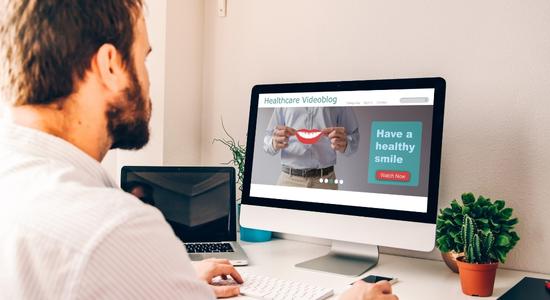 A bearded, brunet man looking at a healthcare videoblog on a screen.