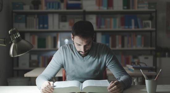 A man reading a very thick book at a desk in a library.