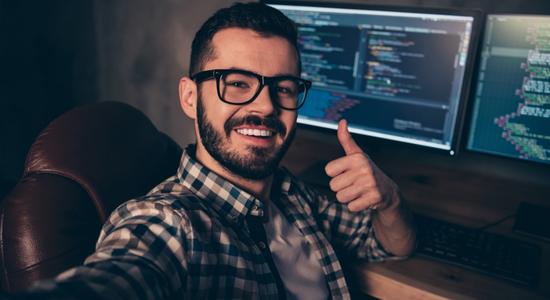 Bespectacled, bearded man smiling, giving thumbs-up in front of a computer. 