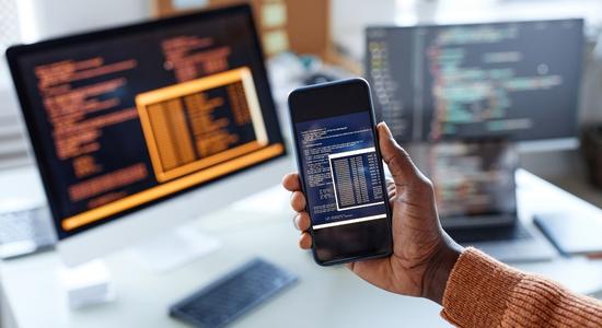 Hand holding smartphone showing tags and strings with computer screen showing the same in the background. 