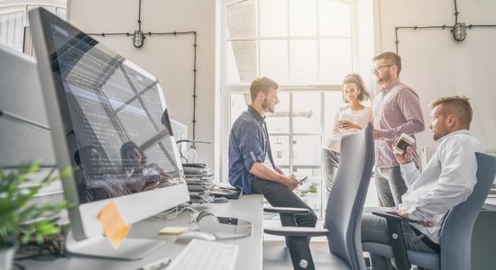 Four people in an office smiling and discussing in sunlight with Mac book screen showing software coding. Concept of software translation services by professional software translators.